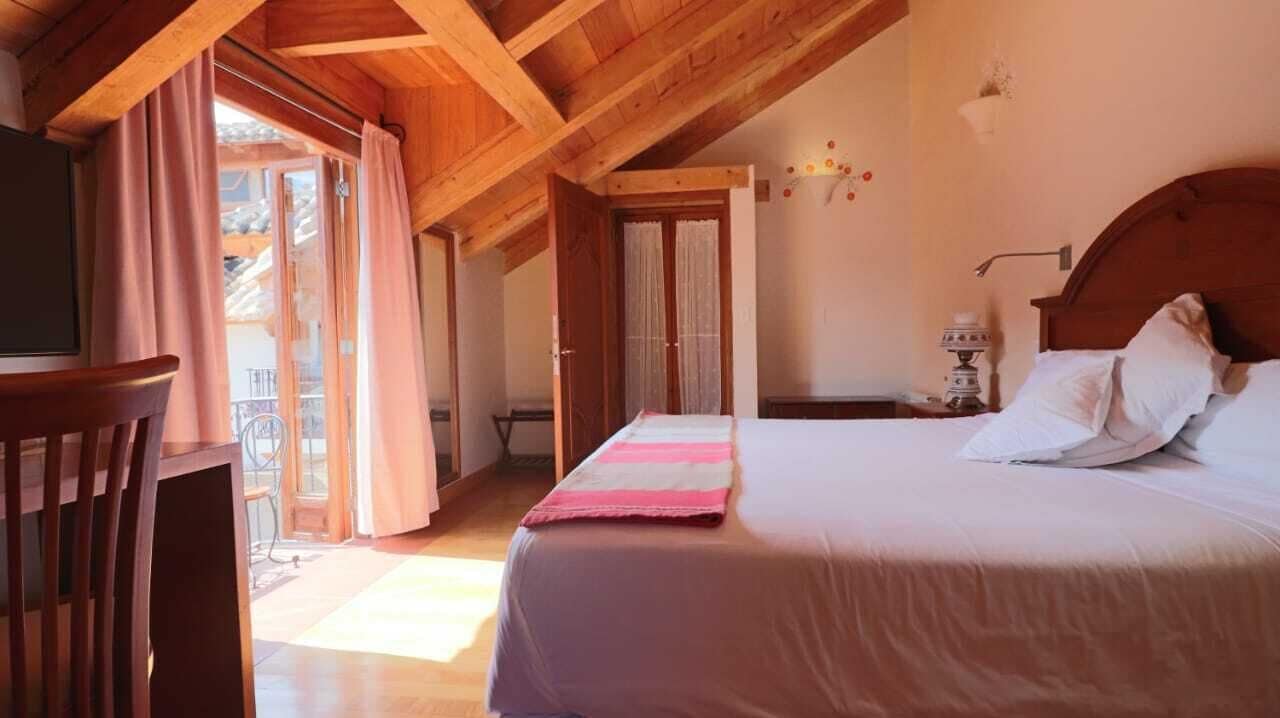 Un dormitorio con techo de madera, una cama con ropa blanca, una manta a rayas rosa y gris, una silla de madera y una ventana con cortinas.