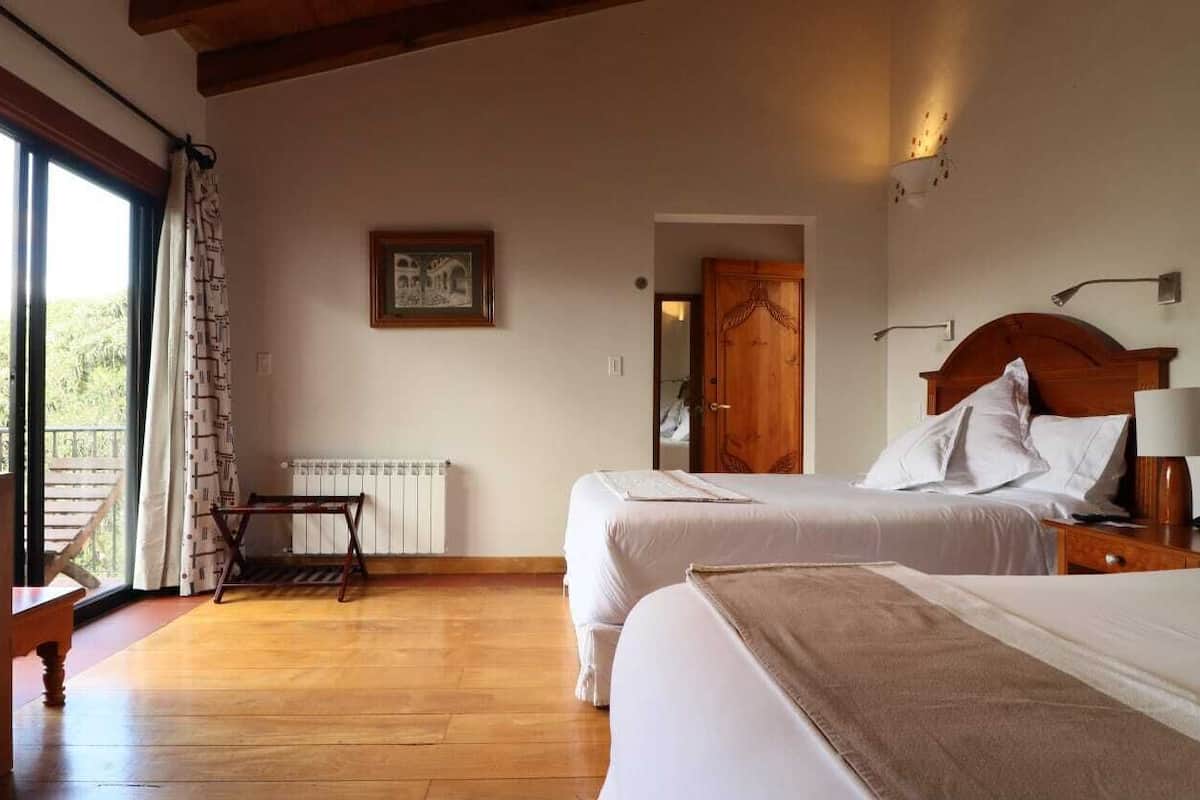 Quarto de hotel com duas camas, piso de madeira, janela com cortinas, radiador e porta que dá para outro cômodo.