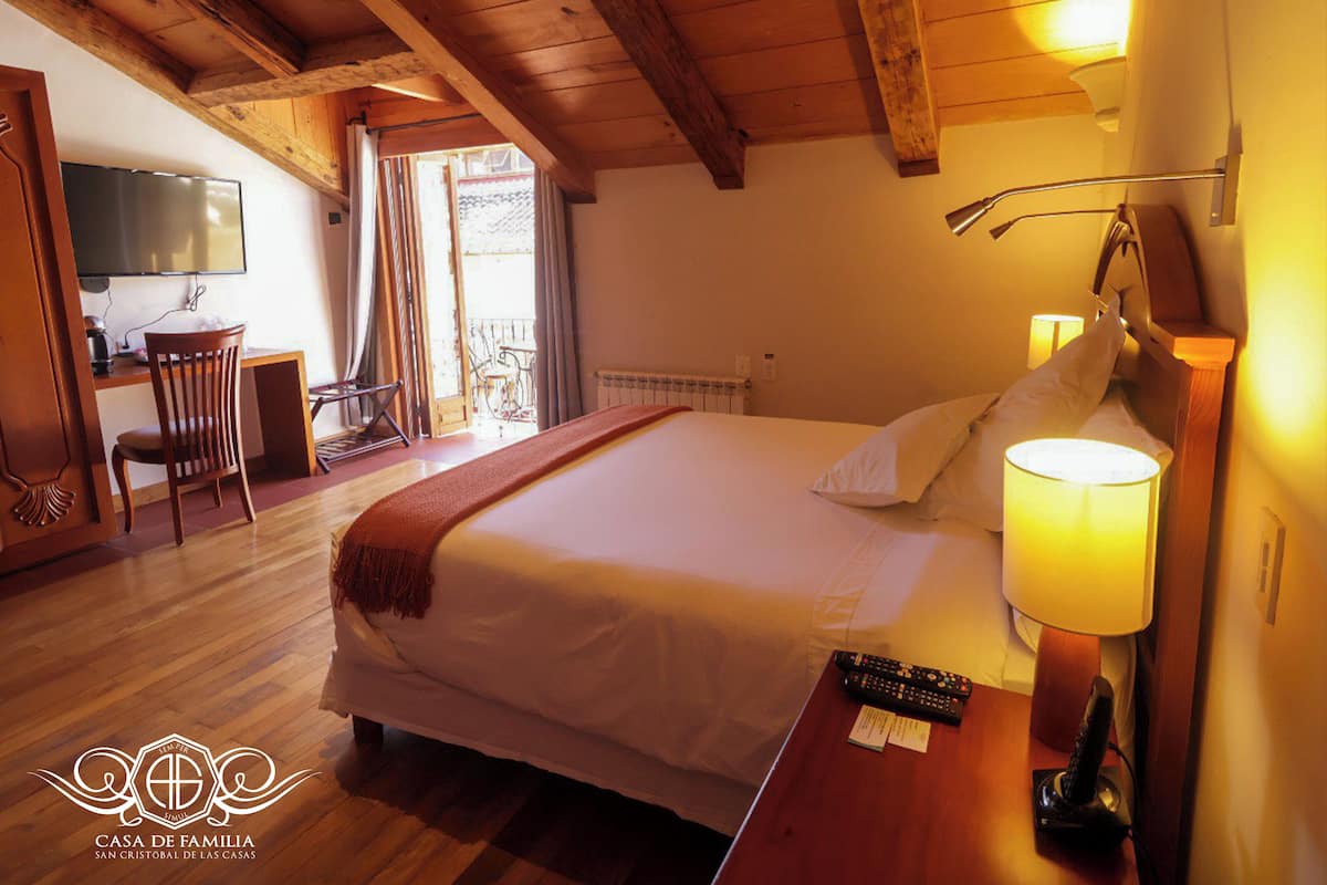 Quarto de hotel com teto de madeira, cama com roupa de cama branca, luminária de cabeceira, televisão e escrivaninha com cadeira.