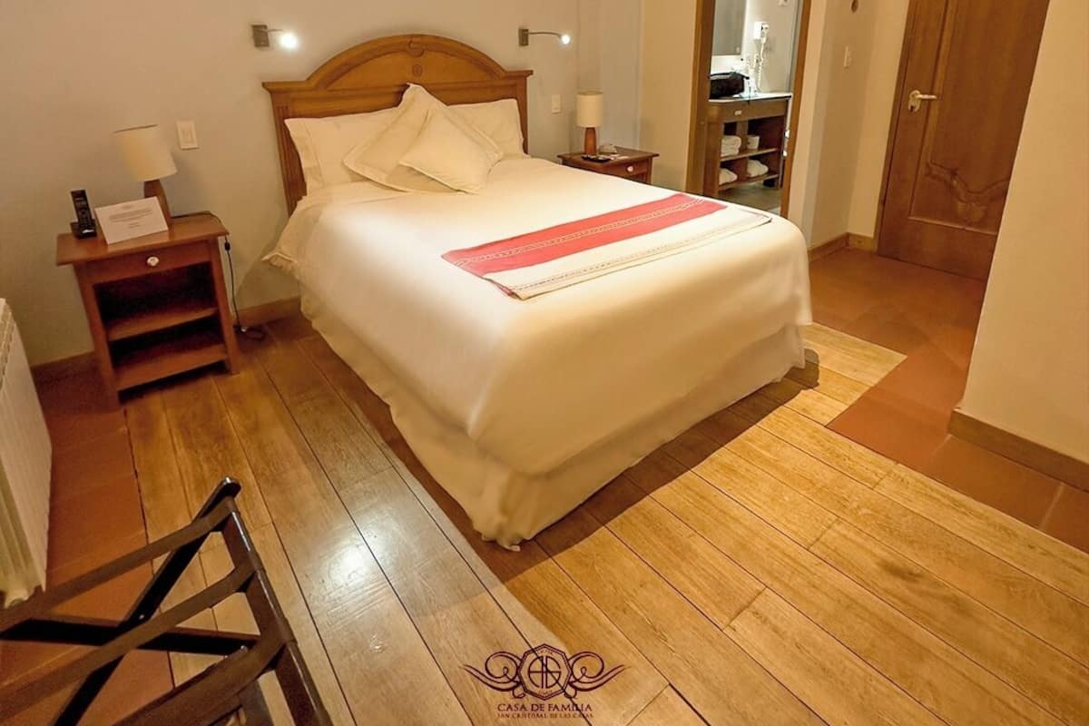 Cama bem arrumada em quarto com assoalho de madeira, cabeceira de madeira e mesa de cabeceira com abajur.