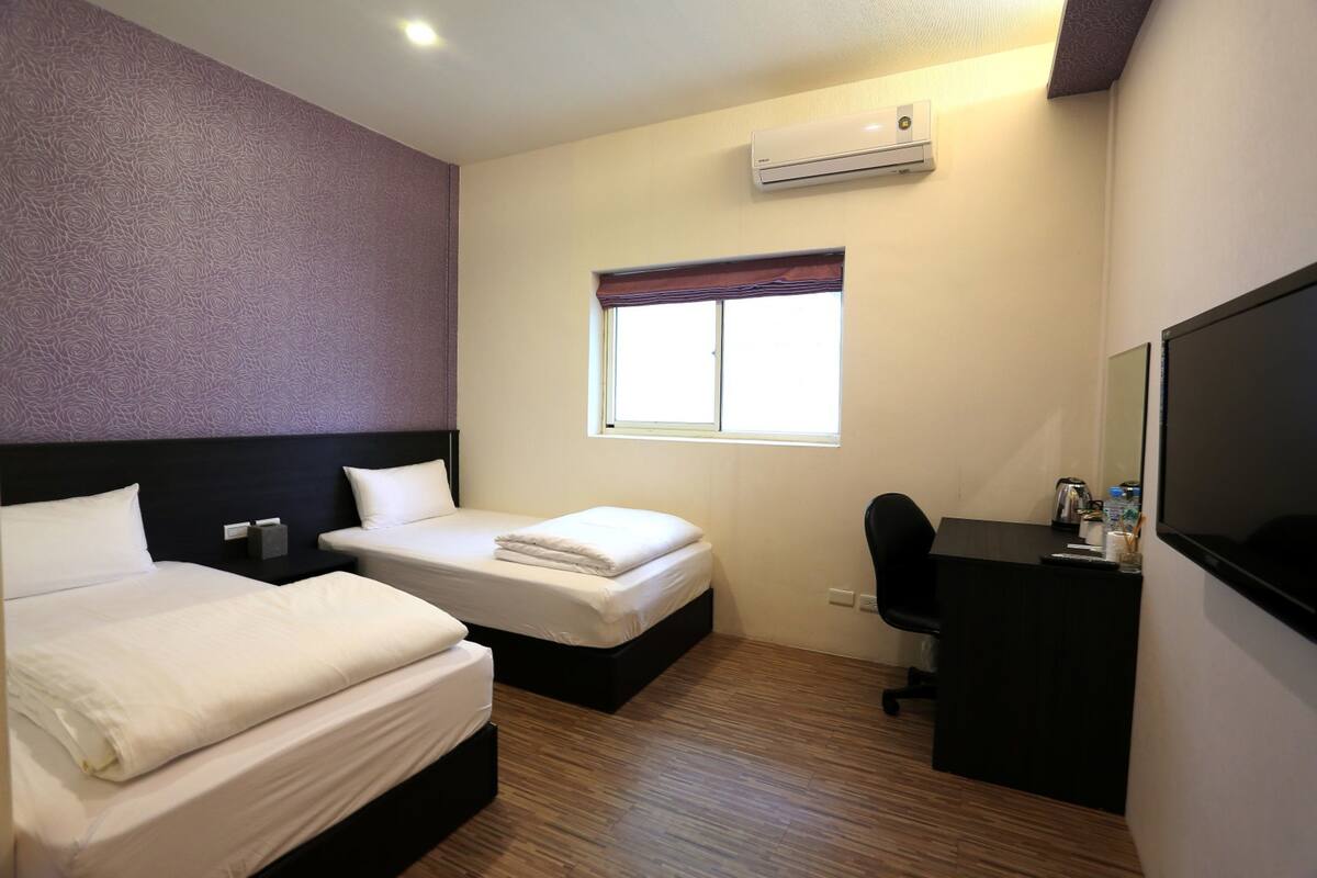 A hotel room with two beds, a desk, a chair, a TV, an air conditioner, and a window with a view.