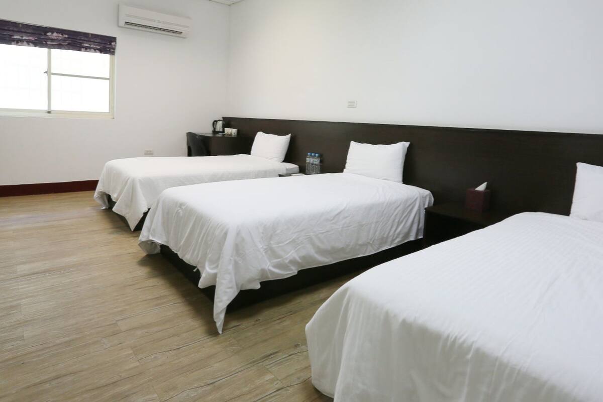 A hotel room with three single beds, a wooden floor, a large window with a view, and a nightstand with a lamp.