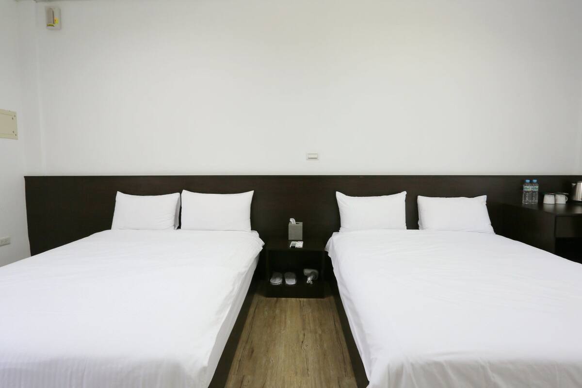 A hotel room with two single beds, a wooden headboard, a small bedside table with a lamp, and a nightstand with a water bottle and a coffee pot.