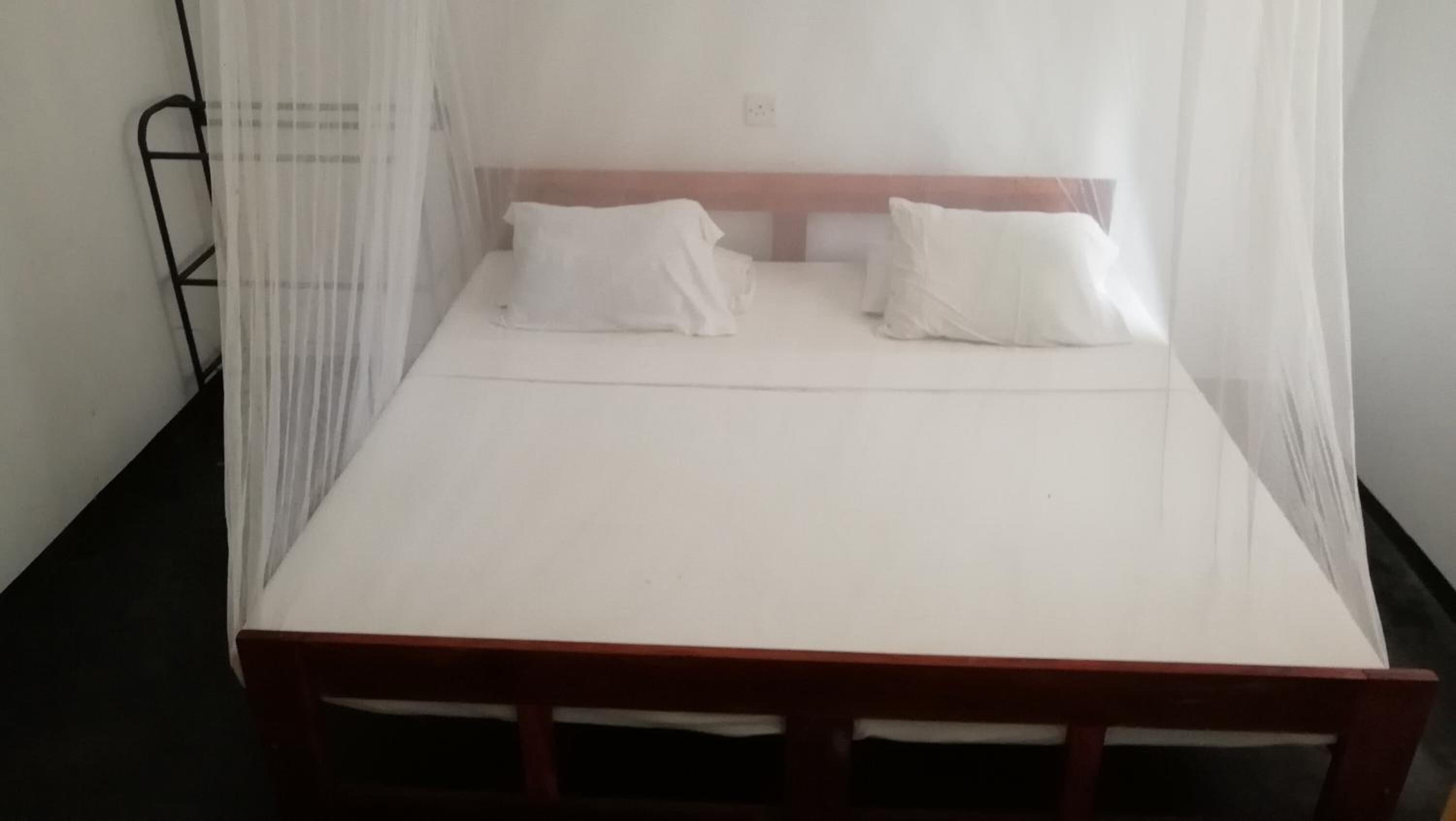 Un llit individual amb estructura de fusta, roba de llit blanca, una mosquitera i una paret sense decoració amb una presa elèctrica.