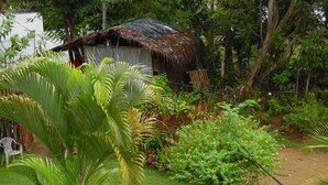 Una cabaña con techo de paja rodeada de exuberante vegetación, entre ellas palmeras y plantas con flores.