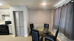 A compact kitchen with a stove, a refrigerator, and a microwave. There is a dining area with a glass table and four chairs. The room has a white wall and a window with curtains.