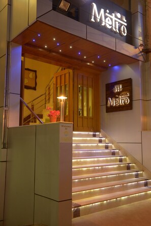 Entrada de hotel con un letrero que dice "Metio", una escalera con escalones iluminados y un cartel de bienvenida.