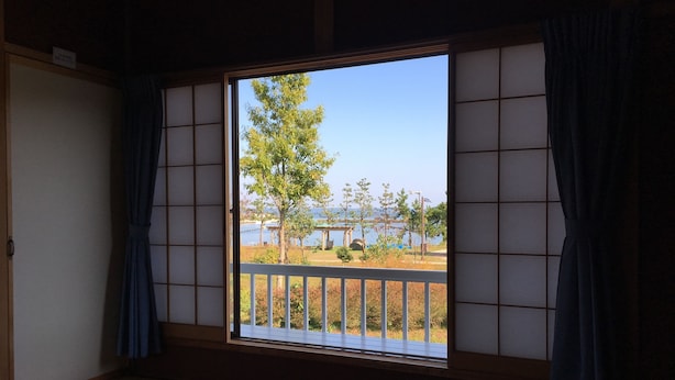 A traditional Japanese room with tatami flooring, a shoji screen, and a view of a park with a tree and a body of water. - Heart Land Hills View of yacht harbor house (Nanao)