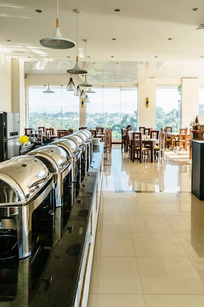 A buffet area with stainless steel appliances and wooden chairs, large windows with a view of greenery, and hanging pendant lights.
