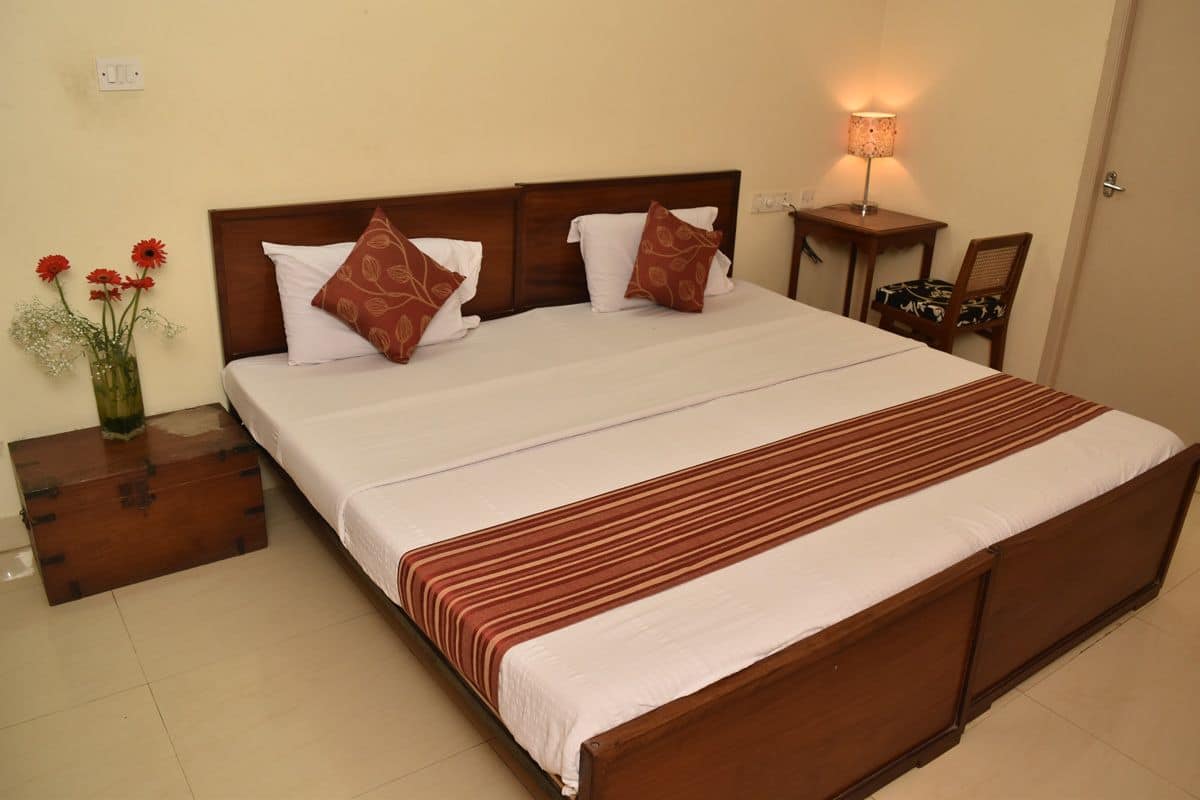 A neatly made bed with a wooden headboard, white bedding, and a striped blanket. There is a small bedside table with a lamp, a chair, and a vase with flowers on the left side.