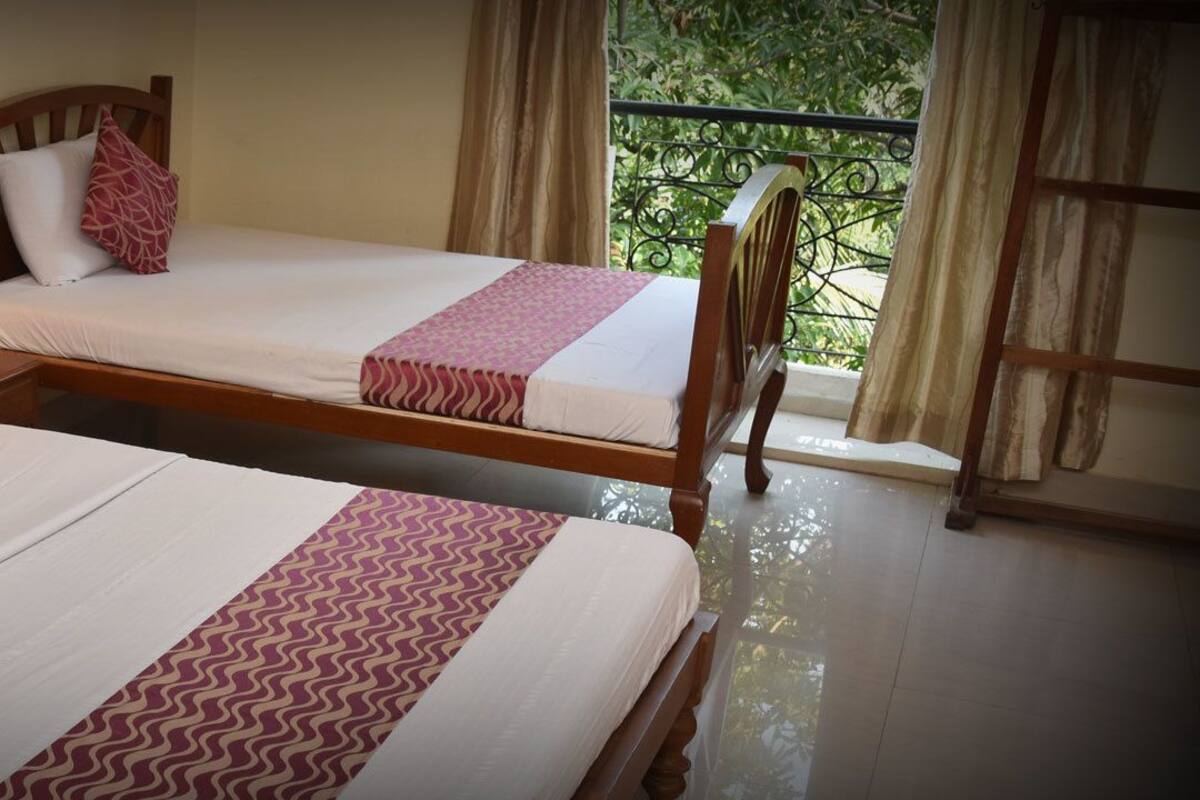 Two adjacent beds with white linens and patterned pillows, a bedside lamp, and a view of greenery through a window.