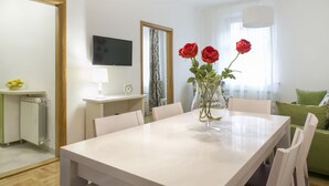 A modern dining area with a white table, four chairs, a vase with red roses, a television, a mirror, and a green sofa. - Apartments Center (Belgrade)