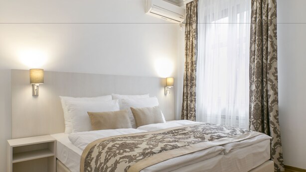 A neatly made bed with a patterned bedspread, two bedside tables with lamps, and sheer curtains. - Apartments Center (Belgrade)
