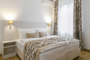 A neatly made bed with a patterned bedspread, two bedside tables with lamps, and sheer curtains on a window. - Apartments Center (Belgrade)