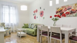 A modern living room with a green sofa, white dining table, and chairs. There is a painting of flowers on the wall and a vase with red flowers on the table. - Apartments Center (Belgrade)