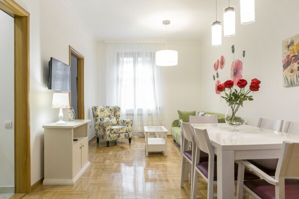 A well-lit room with a dining table, chairs, a small white cabinet, a floral armchair, and a green sofa. - Apartments Center (Belgrade)