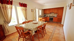 A dining area with a table and chairs, a kitchen in the background, and a window with curtains. - Casa del Campo - sea view villa with private pool in Moraira (Moraira)