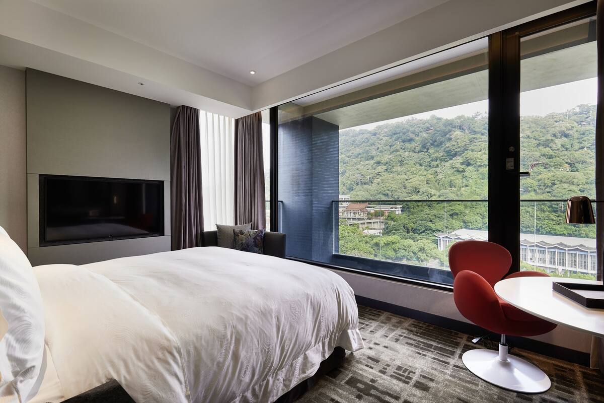 A modern hotel room with a large bed, a TV mounted on the wall, a dining table with a red chair, and a balcony with a view of a forested area.
