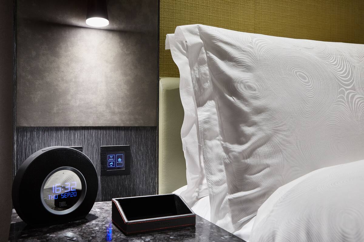 A hotel room with a digital clock, a bedside table, a bed with white bedding, a wall-mounted control panel, and a textured wall.