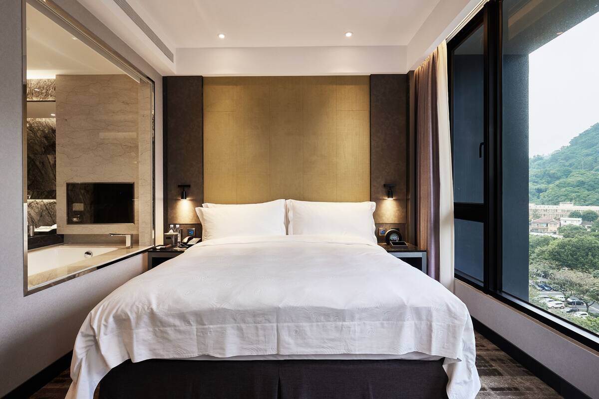A modern hotel room with a large bed, a mirror, a nightstand, and a view of a cityscape through a large window.