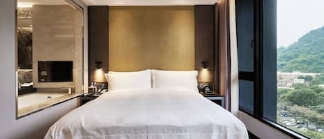 A modern hotel room with a large bed, a mirror, a nightstand, and a view of a cityscape through a large window.