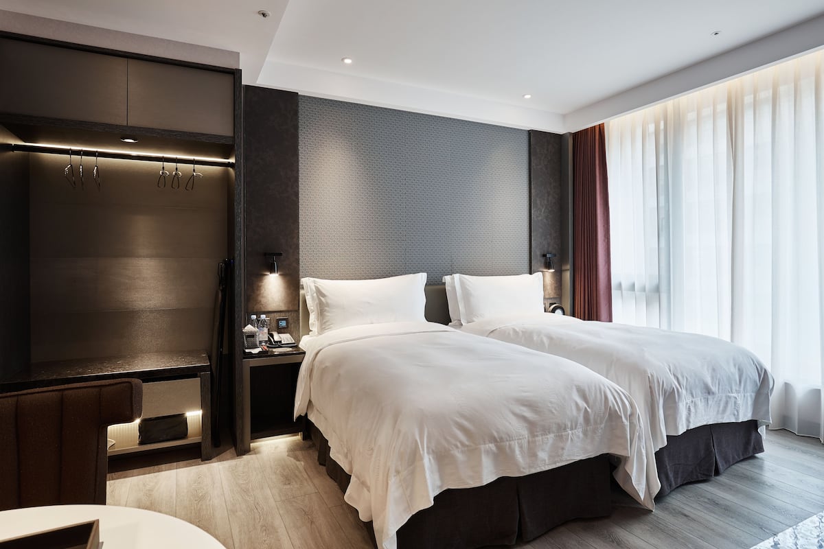 A modern hotel room with two beds, a built-in wardrobe, a bedside table with a lamp, and a large window with sheer curtains.