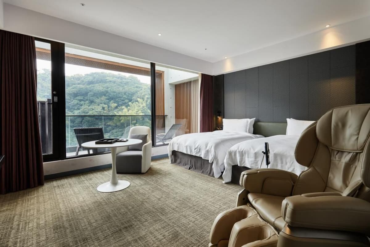A modern hotel room with a large bed, a sofa, a massage chair, and a balcony with a scenic view.