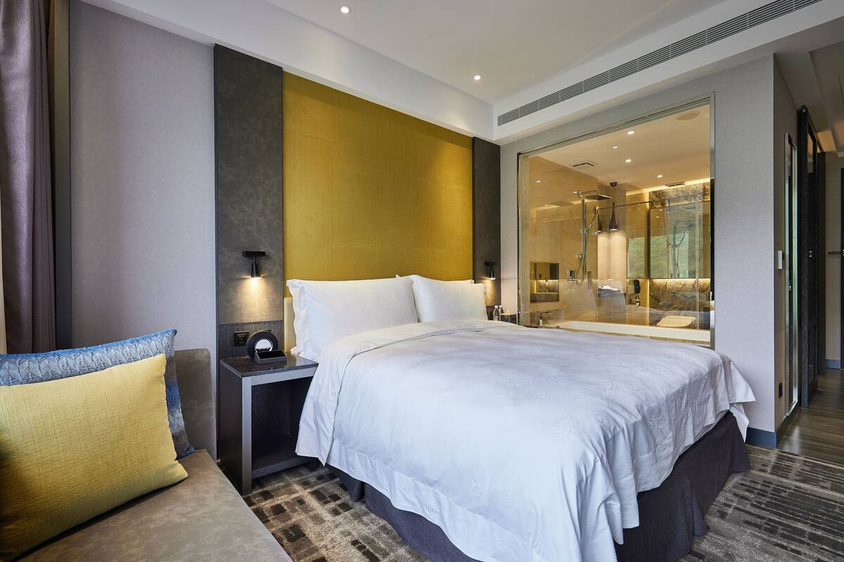 A modern hotel room with a large bed, a bedside table with a lamp, and a glass shower area.