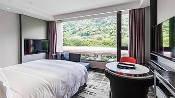 Een hotelkamer met een groot bed, een televisie, een tafeltje en uitzicht op een groen landschap door grote ramen.