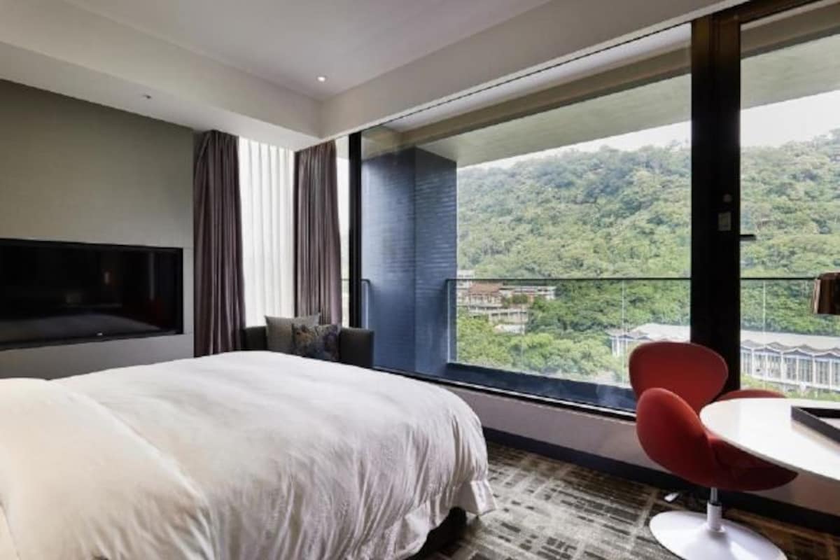 A modern hotel room with a large bed, a TV mounted on the wall, a balcony with a view of a forested area, and a red chair by the window.