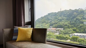 A room with a sofa and a window offering a view of a green hillside and buildings. - The Tango Taipei Jiantan (Taipei)