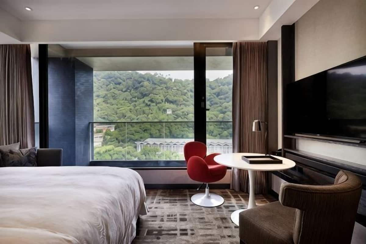 A hotel room with a bed, a red chair, a small table, a TV, and a balcony with a view of a forested area.