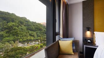 A hotel room with a view of a green hillside, a sofa with pillows, a bedside table with a lamp, and a bed with a white pillow.