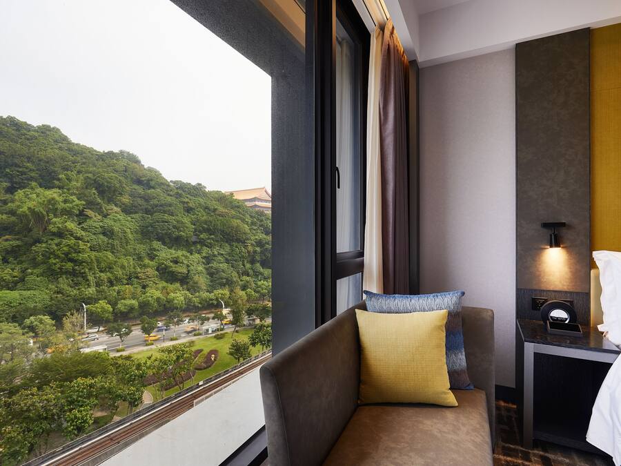 A hotel room with a view of a green hillside, a sofa with pillows, a bedside table with a lamp, and a bed with a white pillow.