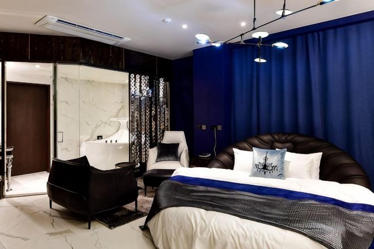 A modern bedroom with a large bed, a sitting area with a chair, a vanity, and a large mirror.