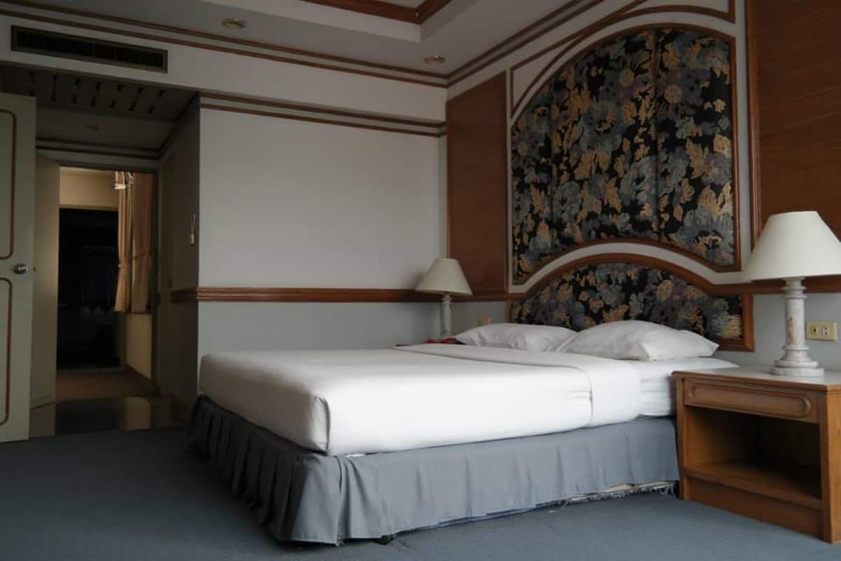Una habitación de hotel con una cama grande, dos mesitas de noche con lámparas, un cabecero estampado con motivos florales y una puerta que da a otra habitación.