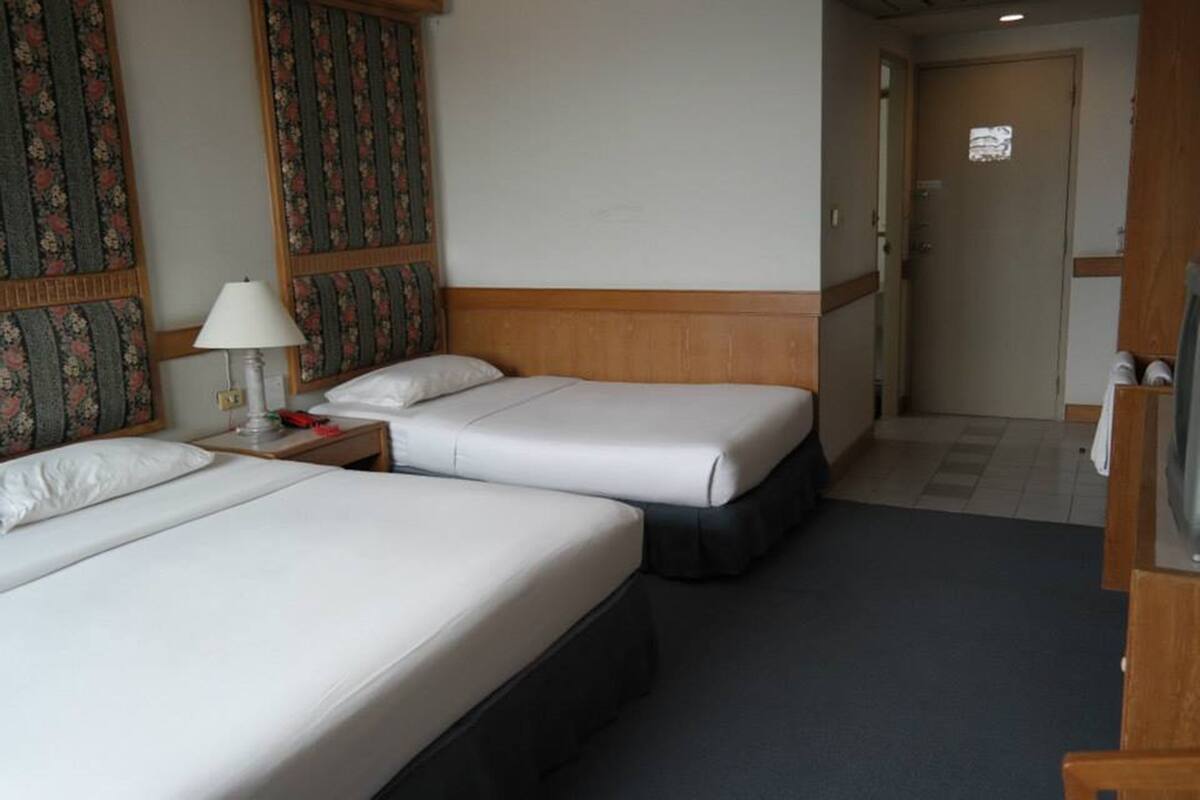 Habitación de hotel con dos camas, una mesita de noche con lámpara, cabecera de madera y una puerta que da a otra habitación.