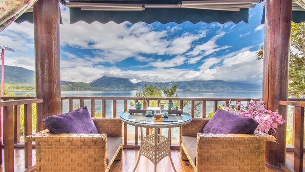 A wooden deck with wicker furniture, a glass table, and a view of mountains and a lake. - Lugu Lake Love Inn (Lijiang)