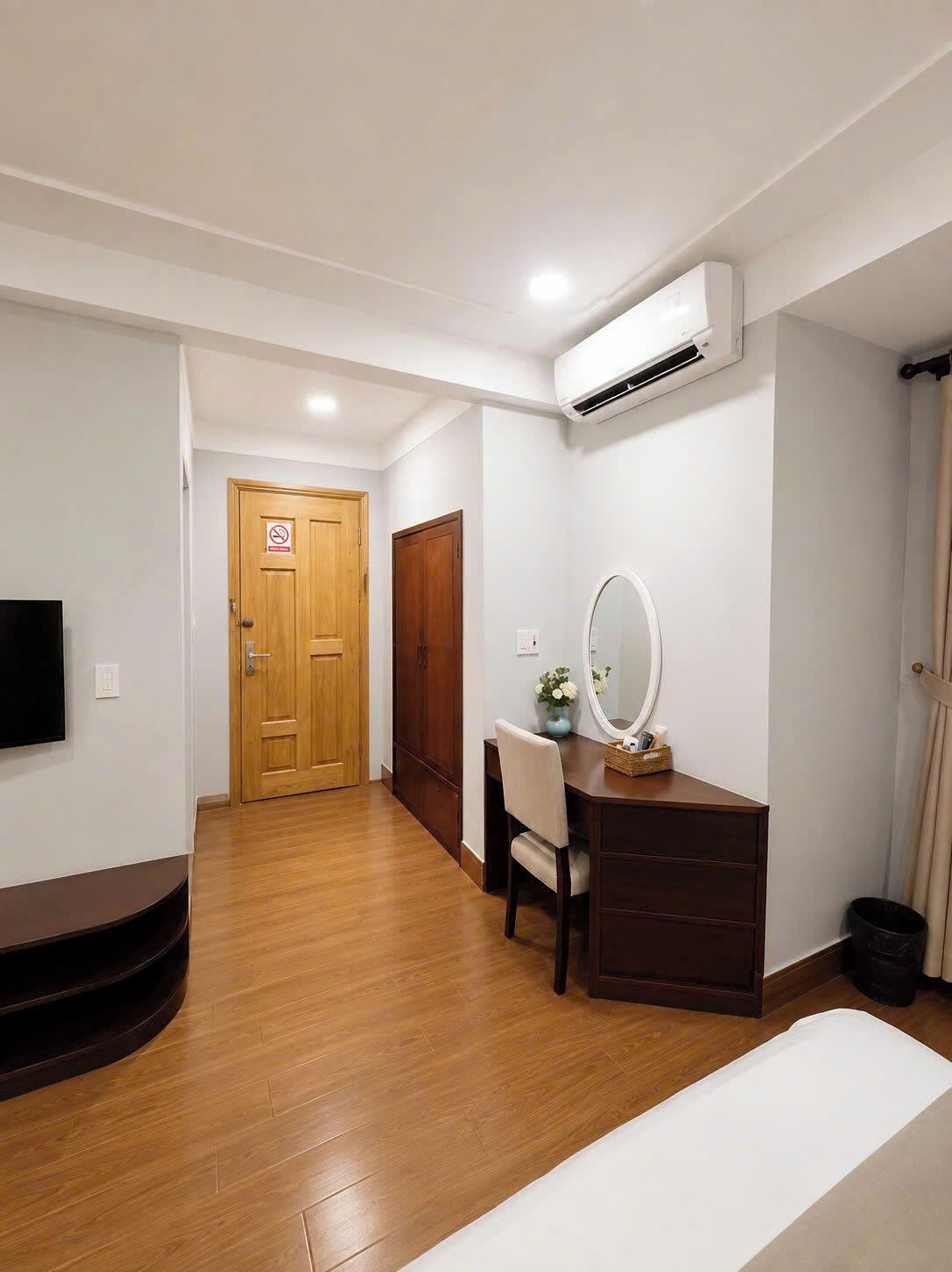A room with a wooden floor, a desk with a mirror, a television mounted on the wall, and an air conditioning unit.