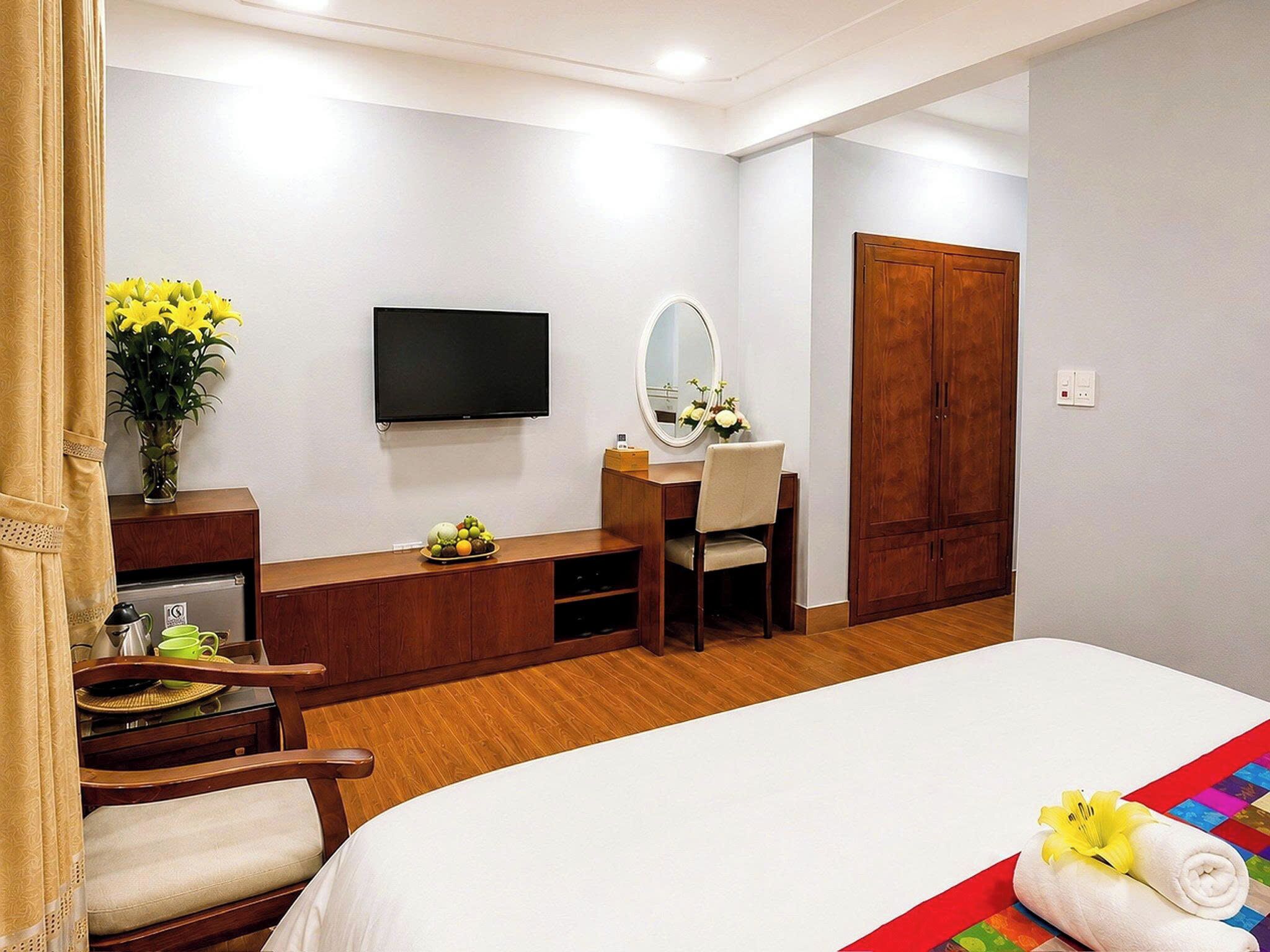 A hotel room with a bed, a desk with a chair, a television, a mirror, a wardrobe, and a vase with flowers.