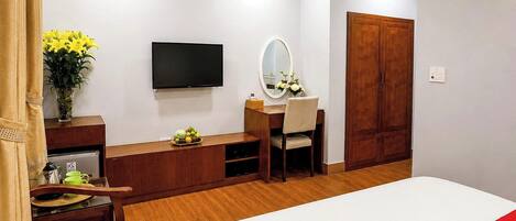 A hotel room with a bed, a desk with a chair, a television, a mirror, a wardrobe, and a vase with flowers.