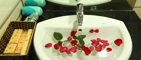 A bathroom sink with rose petals in the water, a towel rack with towels, and a basket of toiletries.