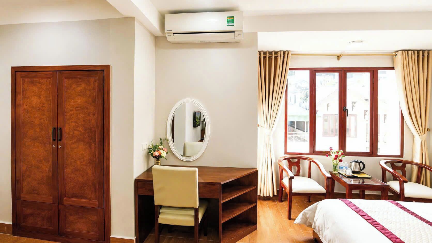 A hotel room with a wooden wardrobe, a desk with a chair, a bed, a window with curtains, and an air conditioning unit.