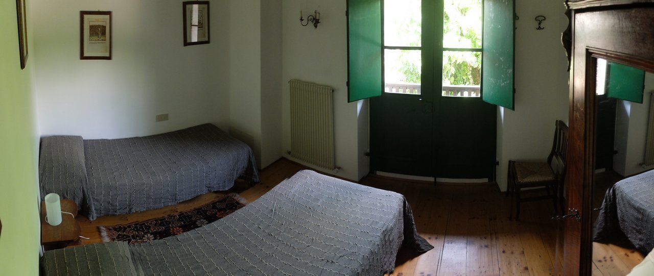 A room with two beds, a radiator, a window with green shutters, a chair, and a door.
