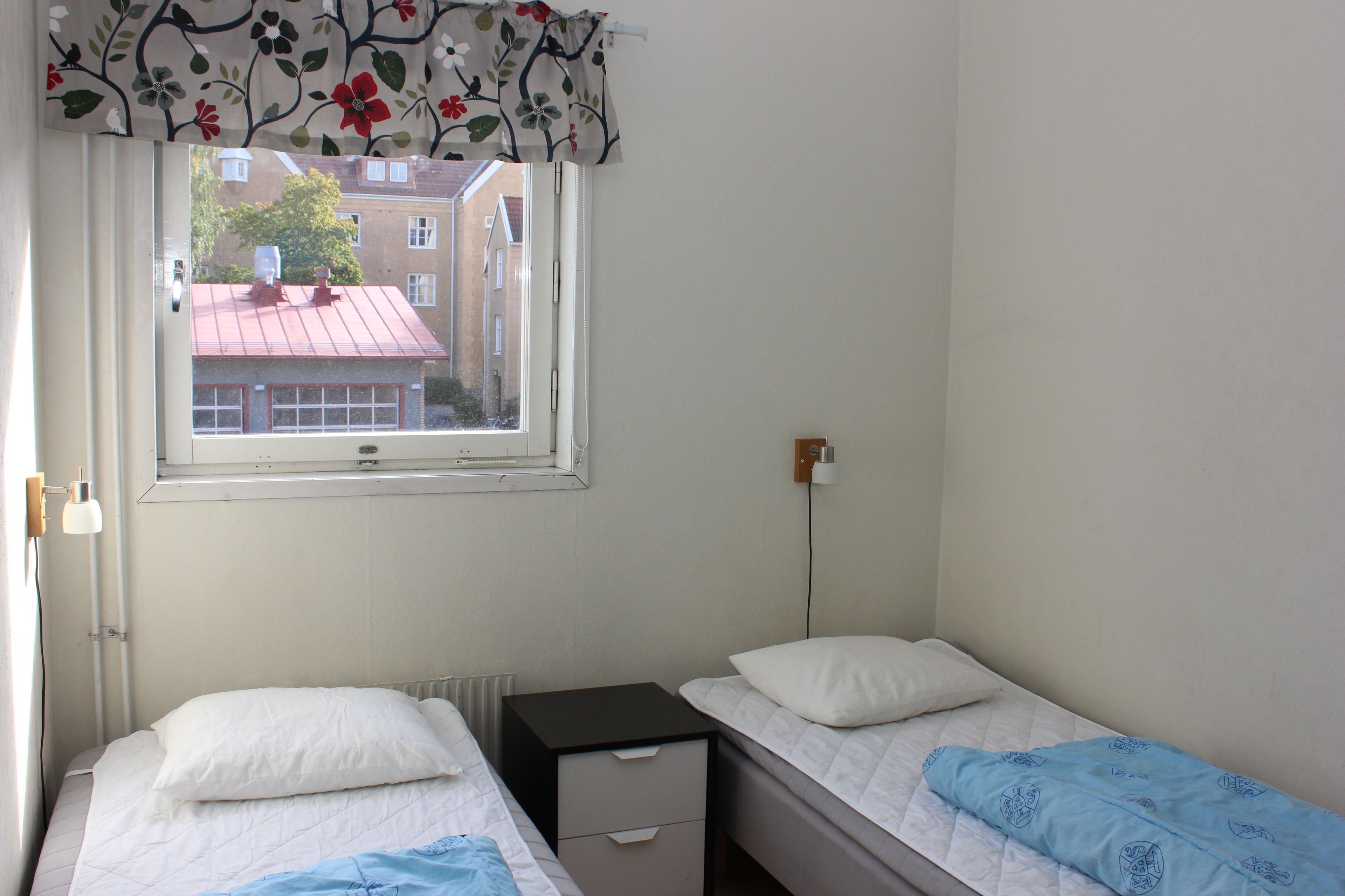 Ett litet, rent rum med två enkelsängar, ett fönster med en blommig gardin, ett element och en väggmonterad lampa.