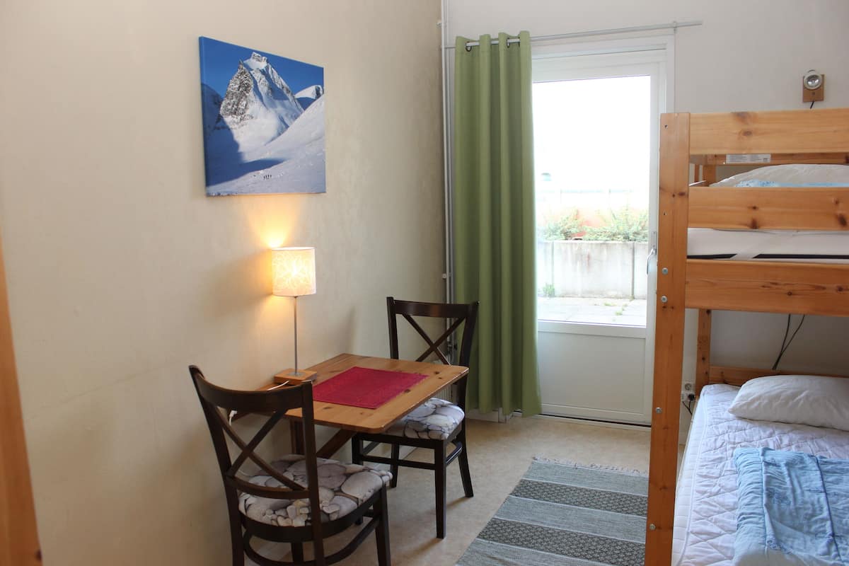 Habitació amb un llit superposat, una taula amb cadires, una làmpada, un quadre i una vista per una porta amb cortines.