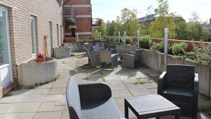 Een terras buiten met rieten meubels, een betonnen plantenbak met bloemen en een bakstenen gebouw op de achtergrond.