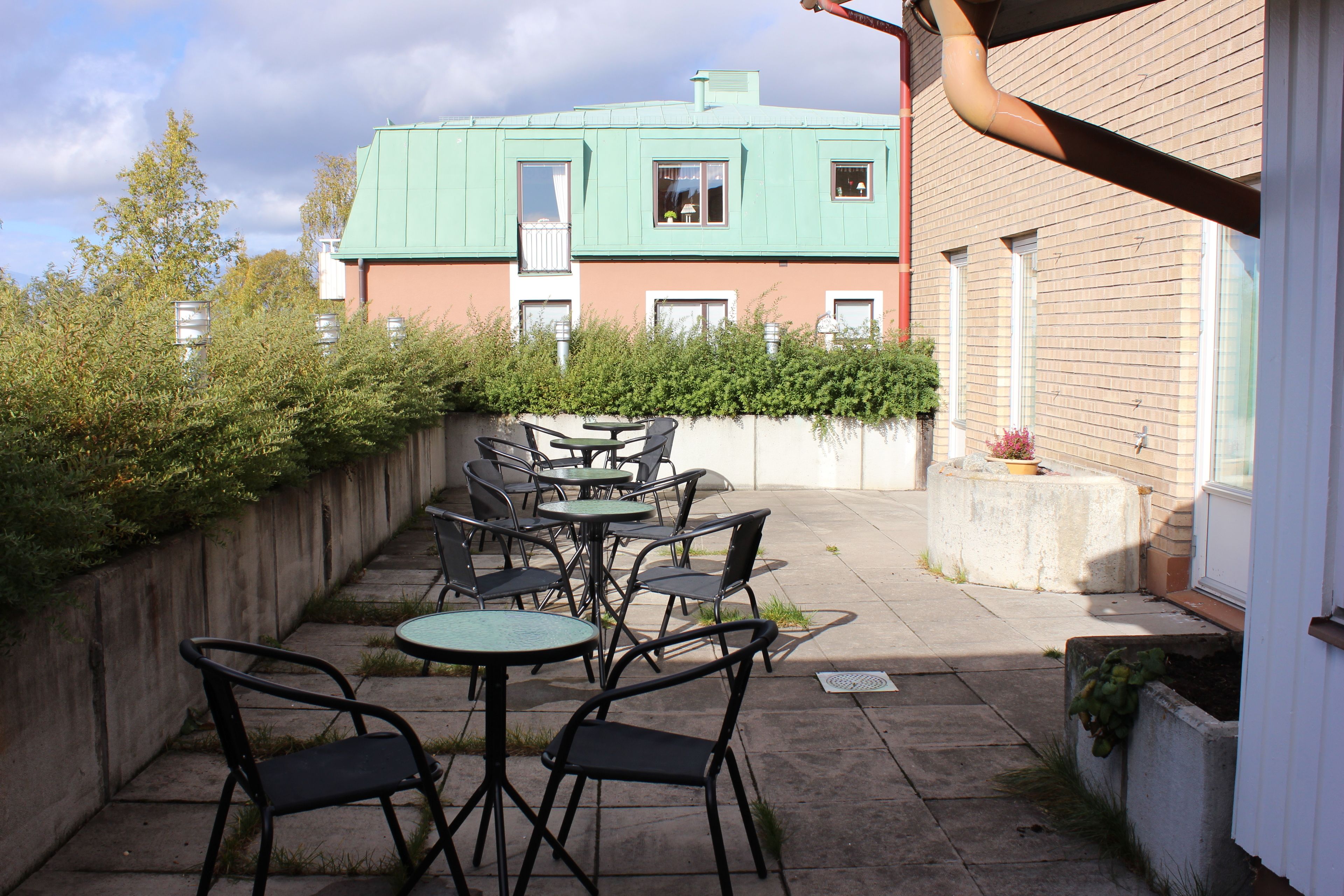 An outdoor patio with tables and chairs, a brick wall, and a building with a green roof in the background.