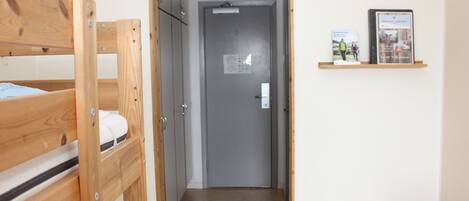 Un pasillo estrecho con una cama litera de madera a la izquierda y una puerta al final. En la pared derecha, hay una estantería con libros y un cuadro enmarcado.