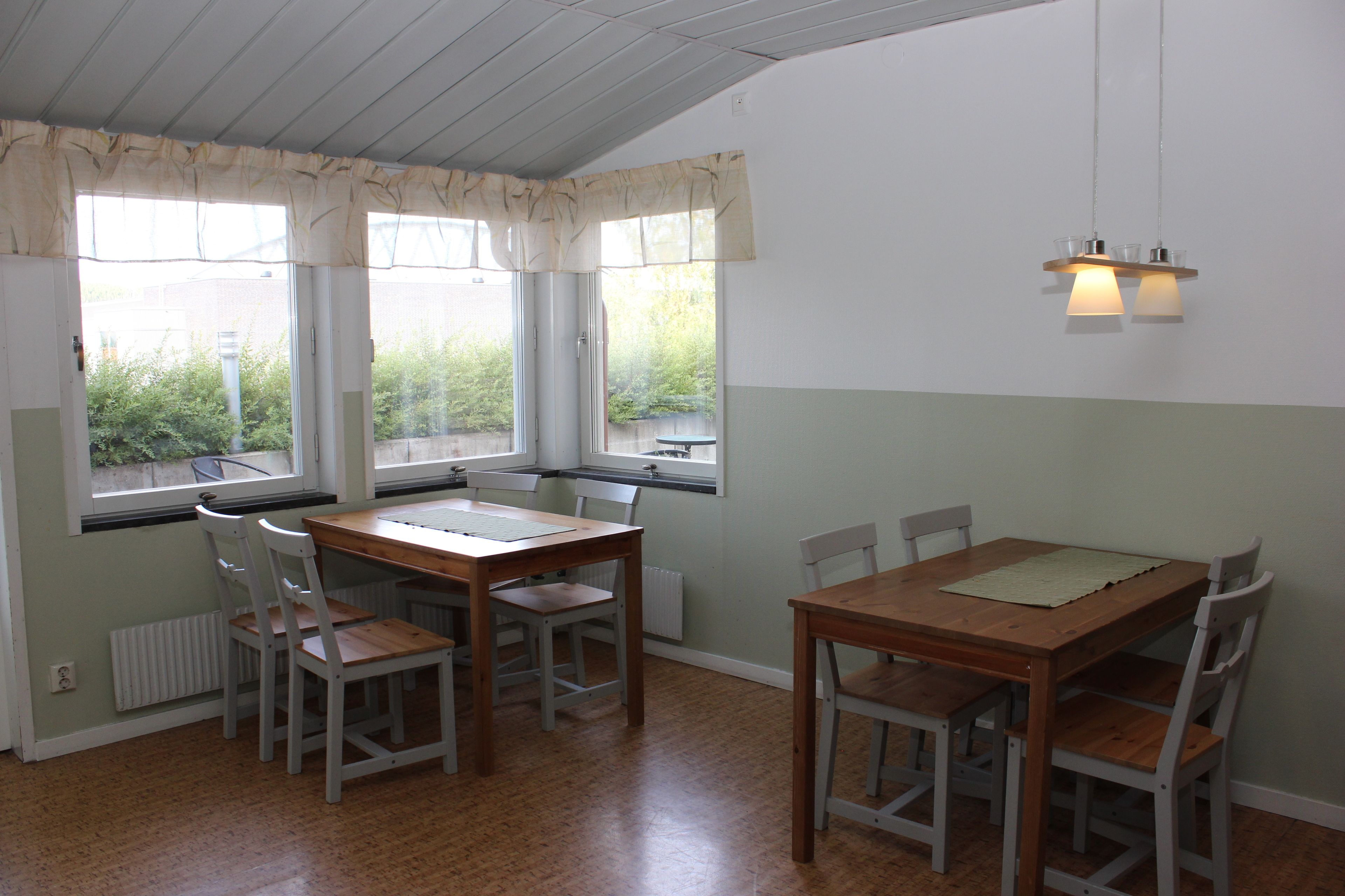 Un comedor con mesa y sillas de madera, ventanas amplias con cortinas y una lámpara colgante.
